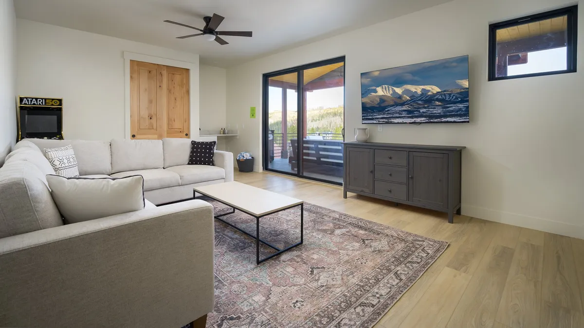 A spacious living room with a comfortable sofa, coffee table, wall-mounted TV, stylish rug, and expansive windows that fill the room with light and scenic views. The arrangement is perfect for game nights, streaming shows, and shared laughter.