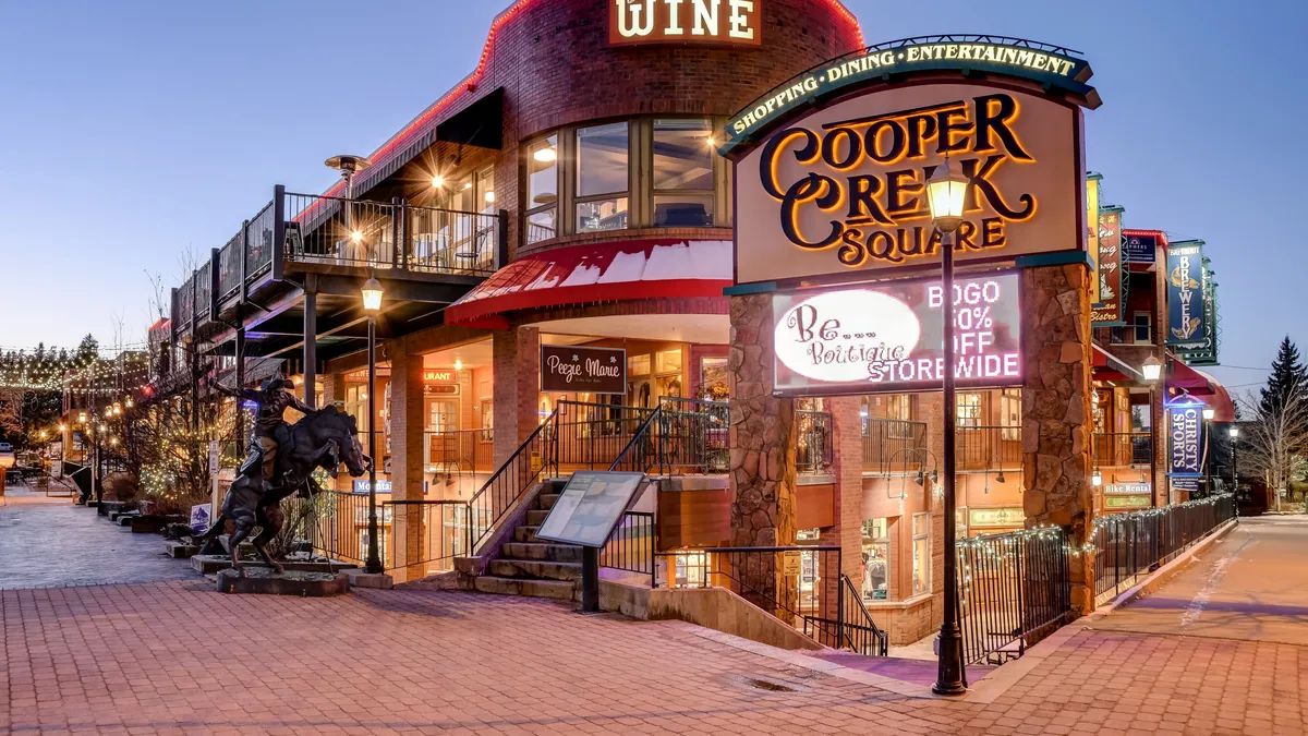 A vibrant outdoor scene featuring the Cooper Creek Square sign, a staircase leading up to a variety of boutique shops, storefronts with colorful signage, and a decorative horse statue in the plaza. People are strolling and enjoying the lively atmosphere.