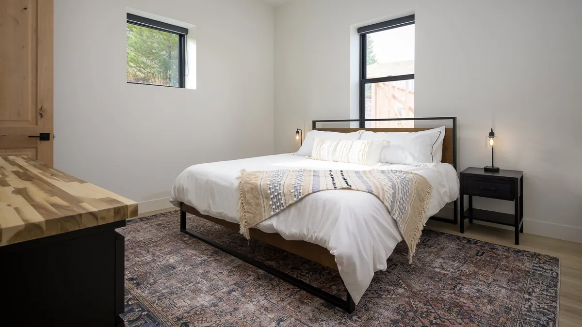 A peaceful bedroom scene featuring a plush bed adorned with a decorative throw blanket, a cozy nightstand with a soft-glowing lamp, and a warm rug beneath. Sunlight streams through the windows, creating a serene and restful ambiance.