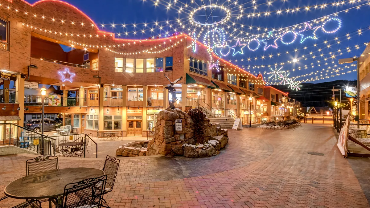 A charming street scene with decorative string lights overhead, shops with large glass windows inviting passersby, and a stone fountain anchoring the plaza. The area is bustling with people enjoying outdoor dining and community events.