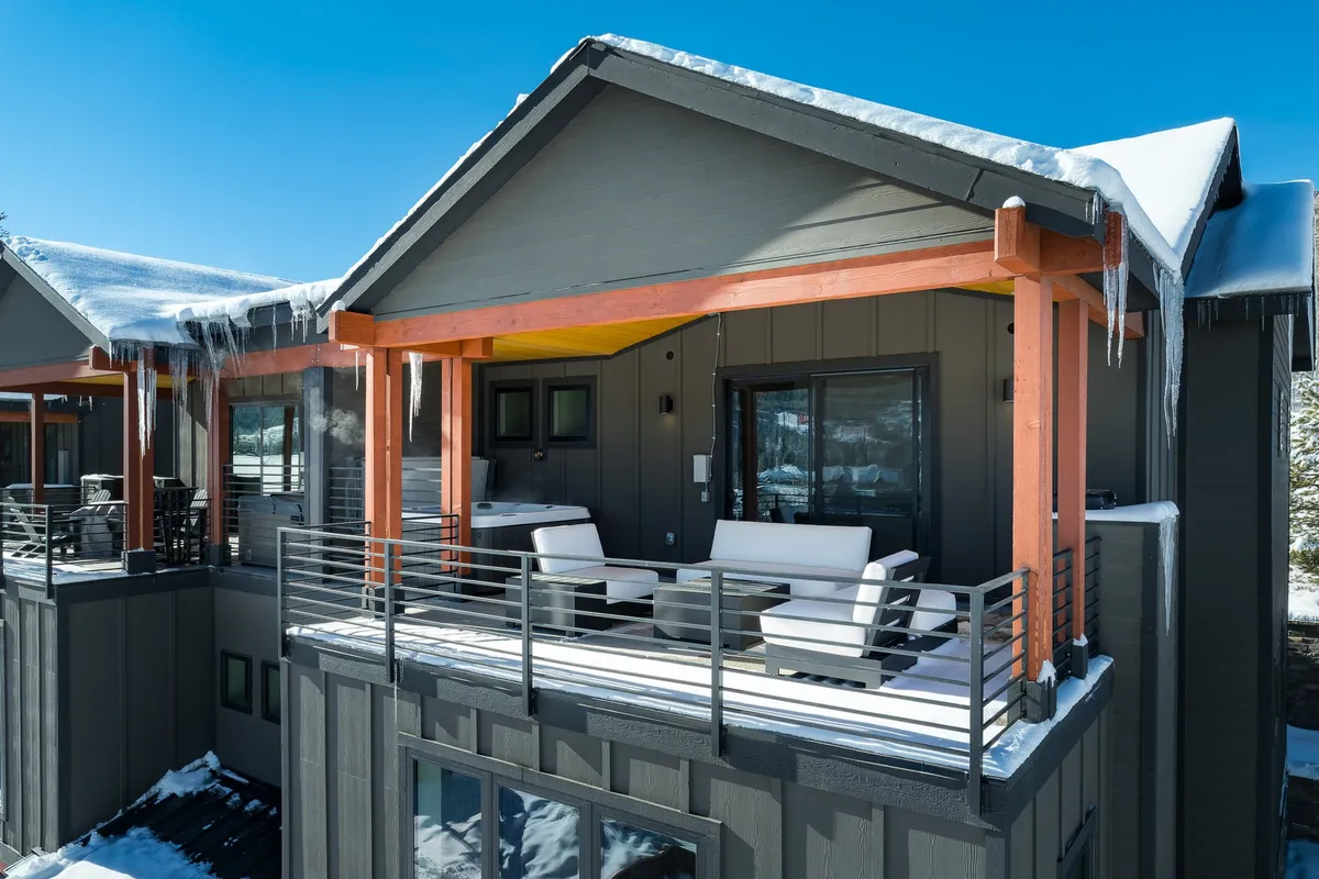 A cozy exterior balcony with inviting seating, orange beams adding warmth to the space, and icicles hanging from the rooftop. The scene looks out over snow-dusted surroundings, suggesting a tranquil vantage point for enjoying mountain views in winter.