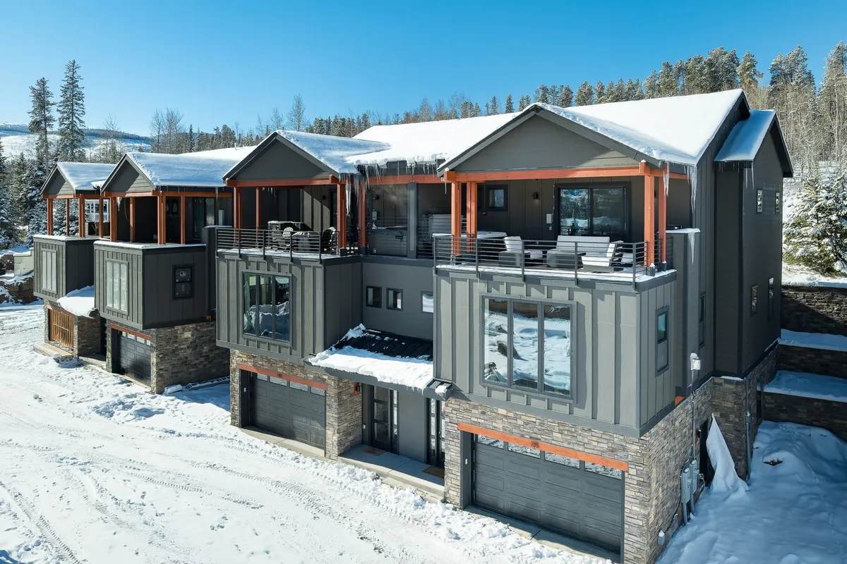 The image showcases a modern, luxurious villa nestled in the snowy landscape of Winter Park, Colorado. The home's striking architecture features a stone facade and multiple balconies dusted with fresh snow, surrounded by tall pine trees. The inviting outdoor spaces hint at relaxing moments with mountain views, while the scene evokes a sense of coziness and tranquility perfect for social gatherings or warming up after winter adventures.