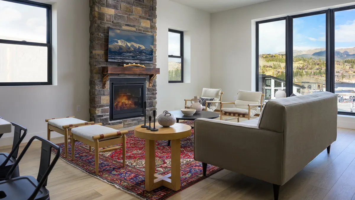 A stylish open living area featuring a prominent stone fireplace, large windows framing mountain views, a cozy rug, and tasteful artwork. The plush seating invites relaxation and lively conversations, all under the warm glow of natural light.